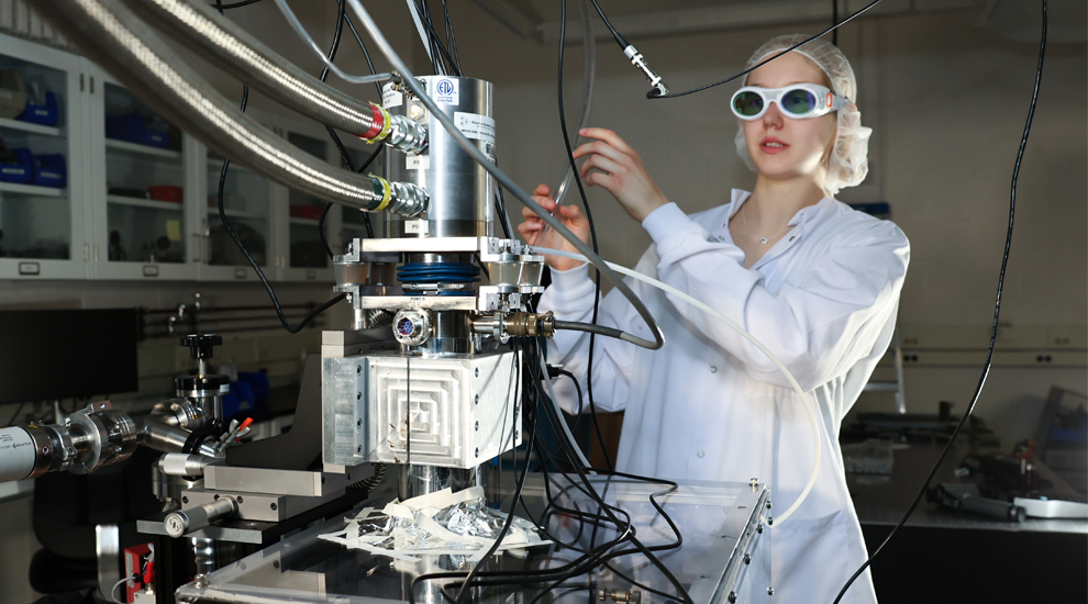 A woman wearing a white lab coat, goggles, and hairnet works on an experiment using lasers in a lab.